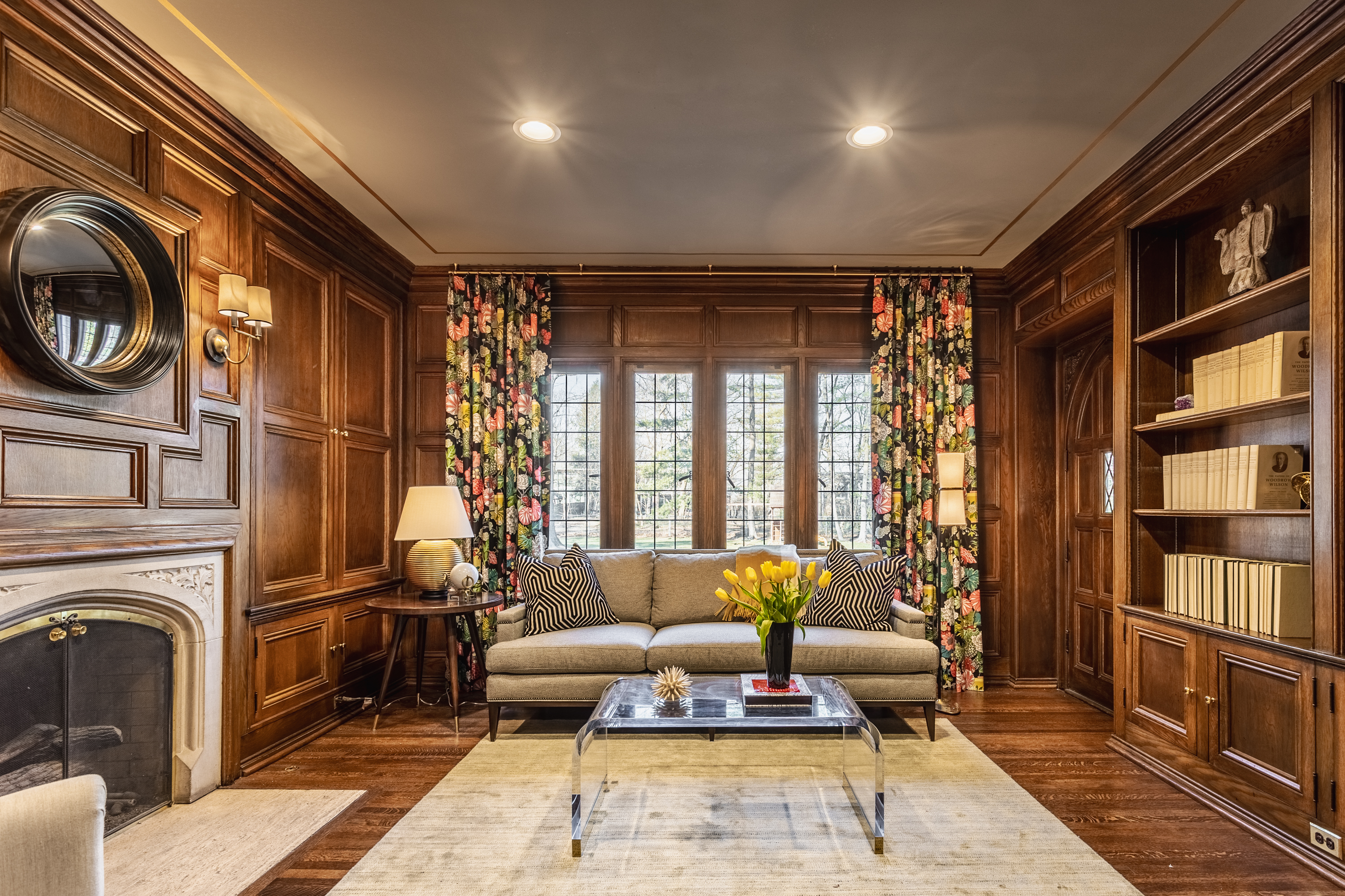 Wood-paneled libraries like this one can easily feel too somber or forbidding. Todd Otterman of Design Works kept the mood stately yet inviting by complementing the traditional millwork and fireplace with Mid-Century Modern panache: the splayed and tapered legs of the side table, the conical shade of the floor lamp (find a similar one here). The transparent waterfall-style coffee table (similar to this one) keeps things light, and the floral curtains are an unexpected splash of traditional femininity in a classically masculine space.
 
 
 
