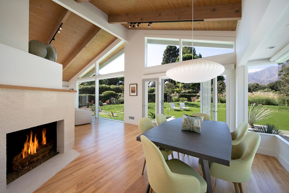 A two-sided fireplace separates the living and dining areas. The green upholstery of the softly curved dining chairs complements the lawn and hedges seen through the windows.
