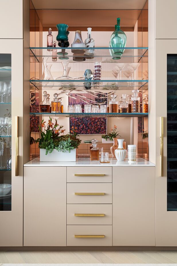 The husband “specifically wanted a beautiful bar cabinet and wine fridge, central to the main area,” Andrew says. The pink and gold tones of the cabinetry, mirror, and hardware ensure that it complements the dining area, where it nestles behind the table.
