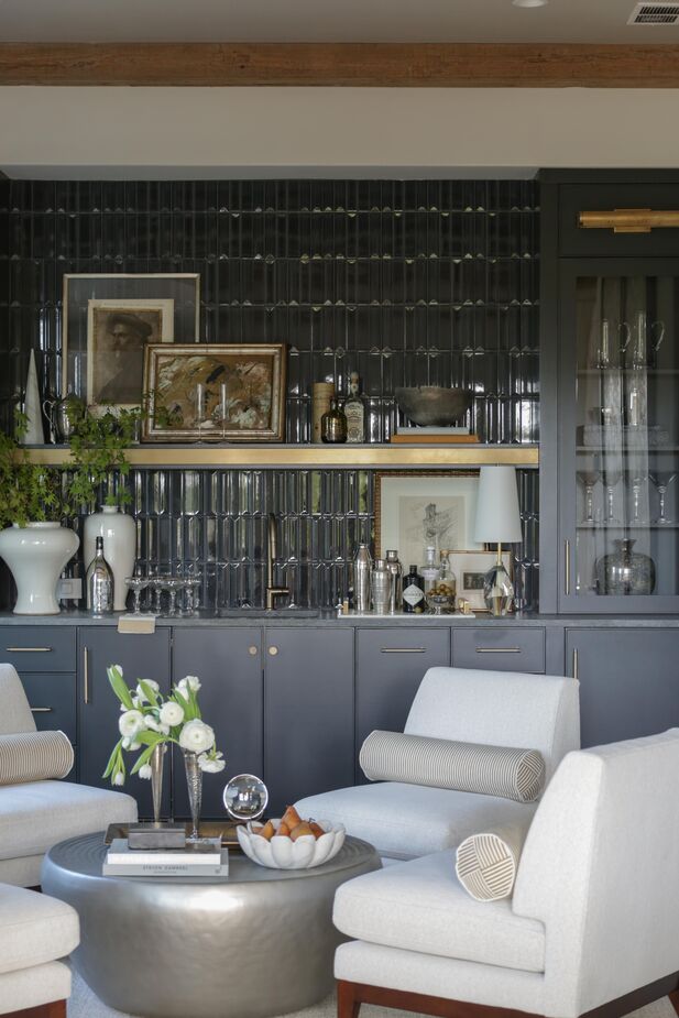 The elongated proportions of this room led designer Ashley Miller to create multiple seating areas. Slipper chairs by the wet bar are ideal for entertaining; “on the opposite side, a backless sofa and two skirted accent chairs create a separate seating area for casual conversation or relaxation,” she explains.

