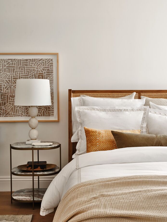 The three tiers of the Freya Oval Nightstand are topped with canyon marble, adding quiet luxury to the minimalist brass frame. Also shown: the Aimee Cane Wingback Bed in Toasted Parawood, the Prado Duvet Cover, and the Chatham Blanket in Linen. 
