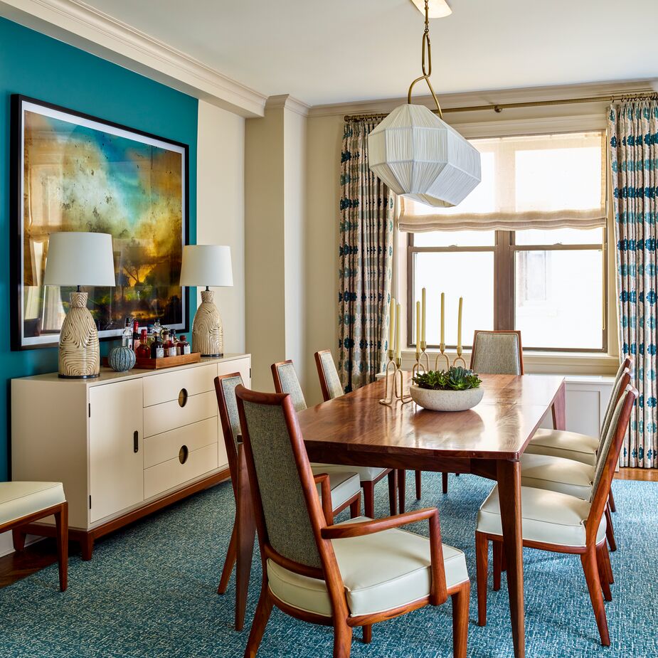 The section of the dining room wall on which a contemporary artwork hangs was painted teal, in contrast to the paler neutral hue of the other walls. This not only spotlights the art but also creates a sense of cohesiveness by tying it in with the teal rug and the similar blues on the curtains.
